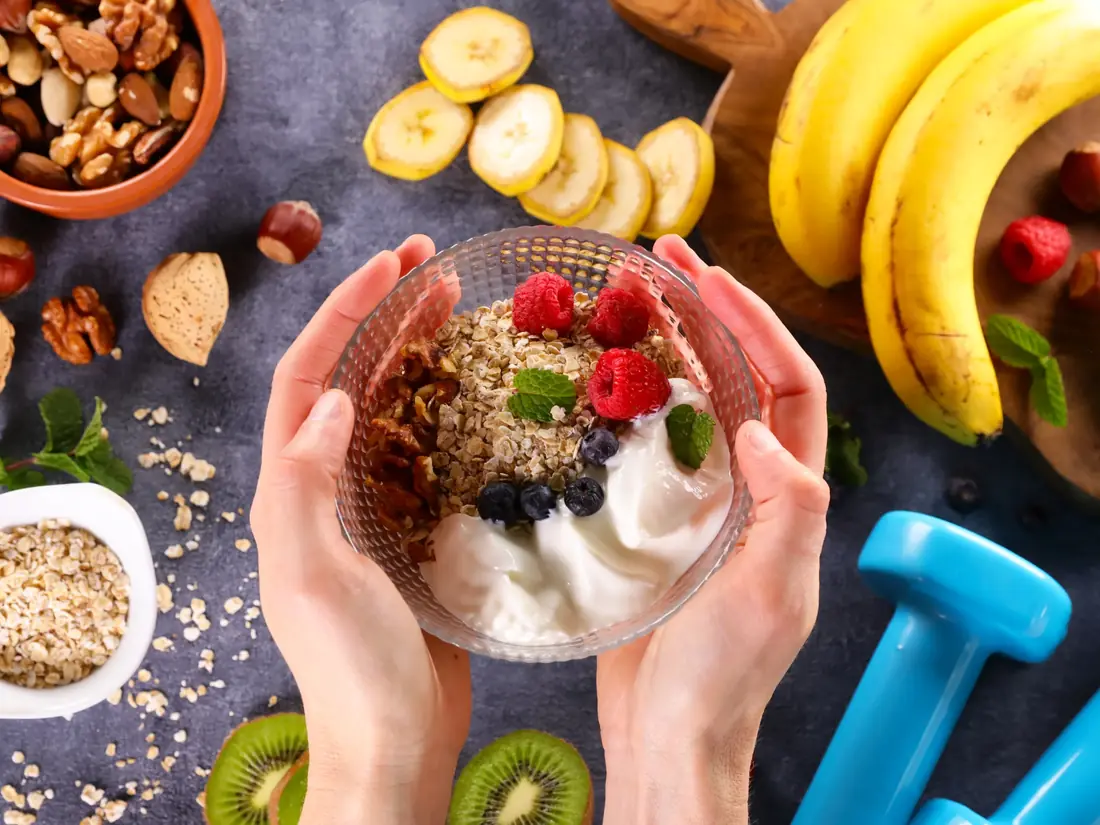 Hands holding a bowl of yogurt, oats, walnuts and fresh berries surrounded by whole foods